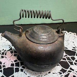 May include: A vintage cast iron teapot with a spring handle. The teapot is dark gray and has a worn, rustic look. It is sitting on a white crocheted doily.