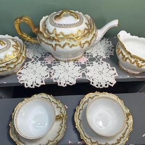 Könnte beinhalten: Ein Vintage-Teeservice mit weißem und goldenem Design. Das Set umfasst eine Teekanne, eine Zuckerdose, einen Sahnekännchen und zwei Tassen mit Untertassen. Die goldenen Verzierungen und Griffe verleihen dem Porzellan-Teeservice einen Hauch von Eleganz.