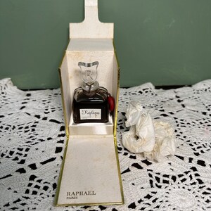 May include: A small, clear glass bottle of perfume with a gold cap and a red tassel. The bottle is labeled "Replique" and "Raphael Paris". The bottle is sitting in a white box with a gold trim.
