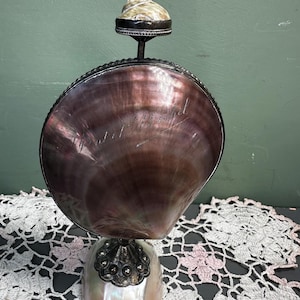Puede incluir: Una escultura decorativa de concha con una gran concha iridiscente de color púrpura oscuro como cuerpo principal. La concha está montada sobre una base nacarada y rematada con una concha más pequeña. La escultura tiene adornos y detalles metálicos plateados.