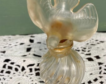 May include: A vintage clear glass perfume bottle with a bird-shaped stopper. The bottle has a spiral design and a slightly aged appearance. The bird stopper is detailed with wing and feather features.