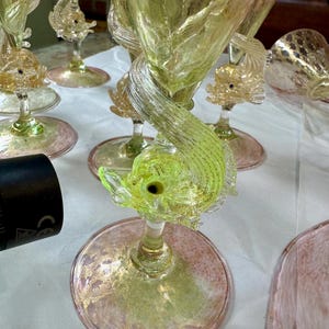 May include: A collection of ornate, hand-blown glass goblets. Each goblet features a fish-shaped accent and a twisted stem. The base and bowl have a pink and gold speckled design, with a light green and yellow color scheme.