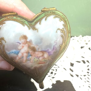 Puede incluir: Una caja de porcelana para alhajas en forma de corazón con un exterior de esmalte verde y detalles dorados. La tapa presenta una escena pintada de dos querubines jugando con un arco y una flecha.