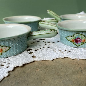 May include: Set of four small, light blue ceramic bowls with handles. The bowls have a textured, dotted surface and are decorated with a floral design. The handles are light green and adorned with small white dots. The bowls are displayed on a white lace doily.