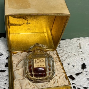 May include: Vintage perfume bottle labeled "JOYA MYRURGIA" in a gold-colored box. The perfume bottle is clear with a geometric design and contains a dark liquid. The box is lined with cream-colored fabric.