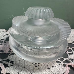 Può includere: Un flacone di profumo in vetro trasparente con finitura satinata. Il flacone ha una base rotonda e una parte superiore decorativa a strati con un design a coste. Anche il coperchio è a coste. Il flacone poggia su un centrino di pizzo bianco.