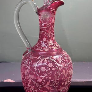 May include: A ruby red glass decanter with a clear glass handle and stopper. The decanter is adorned with white floral and vine designs. The base is a clear, ornate, circular design. The decanter is a decorative piece.