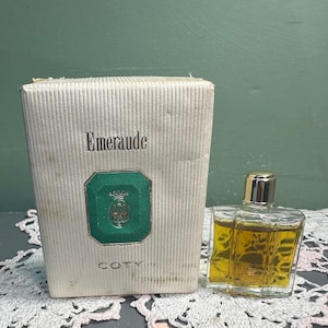 Può includere: Flacone di profumo "Emeraude" vintage e scatola. La scatola è bianco sporco con linee verticali e la parola "Emeraude" in nero. Un emblema verde e oro è presente sul davanti. Il flacone è trasparente e contiene un liquido giallo.