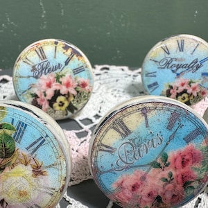 May include: A collection of round decorative objects with a vintage clock face design. Each piece features Roman numerals, floral accents, and text such as "Fleur," "Royalty," and "Paris" on a light blue background.