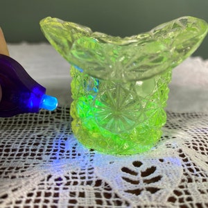 May include: A small, light green glass top hat-shaped container with a diamond pattern. The glass glows bright green under a UV light. The hat sits on a white lace surface.
