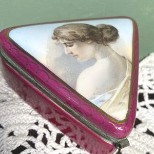 May include: A triangular, antique trinket box with a hinged lid. The box is a deep magenta color with gold trim. The lid features a painted portrait of a woman in profile, wearing a pearl necklace. The background is a pale blue.