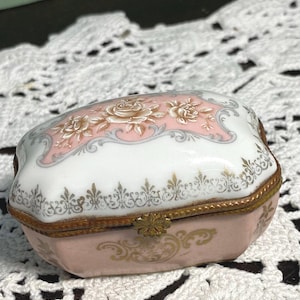 May include: A small, antique porcelain trinket box. The box is primarily pink and white, with a floral design on the lid. Gold accents are present on the edges and clasp. The box is likely used for storing jewelry or small keepsakes.