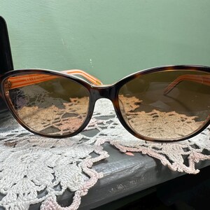 Puede incluir: Gafas de sol de ojo de gato con montura carey y lentes degradadas. Las patillas son de color naranja claro. Las gafas de sol est&aacute;n sobre una superficie de encaje decorativo, con un estuche negro en el fondo.