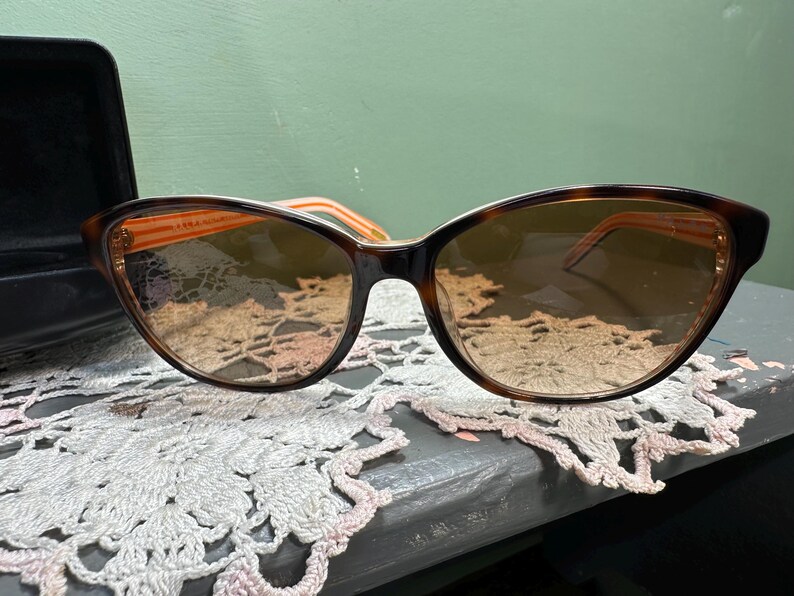 Puede incluir: Gafas de sol de ojo de gato con montura carey y lentes degradadas. Las patillas son de color naranja claro. Las gafas de sol est&aacute;n sobre una superficie de encaje decorativo, con un estuche negro en el fondo.