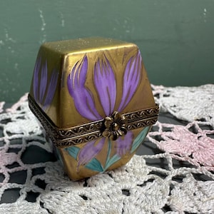 May include: A gold-colored, hexagonal trinket box with a hinged lid. The box is decorated with purple floral designs and a bronze-colored flower clasp. The base of the box has a turquoise and gold painted design. The box is sitting on a white lace doily.
