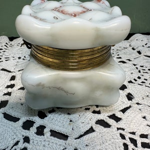 May include: A white porcelain trinket box with a hinged lid. The lid has a raised, scalloped design with pink accents. The box has a brass hinge and a brass band around the bottom.