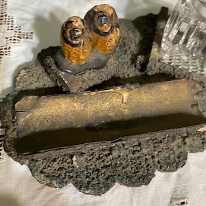 May include: A vintage inkwell set with a black and brown ceramic owl design. The inkwell is made of a textured, dark gray material and has a rectangular shape. The set includes a glass inkwell.