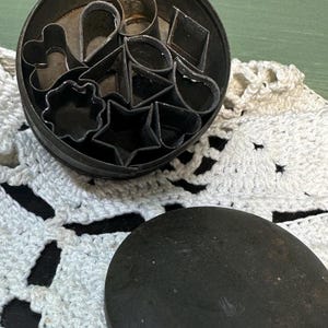 May include: A round metal tin filled with various cookie cutters in different shapes, including hearts, stars, and leaves. The tin is open and the lid is sitting next to it on a white crocheted doily.
