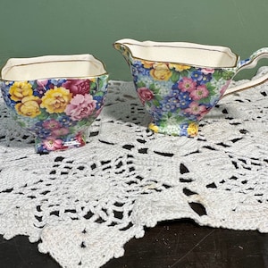 May include: A vintage floral patterned sugar bowl and creamer set. The set is made of white porcelain with a gold trim and features a colorful floral design. The sugar bowl and creamer are both on a white crocheted doily.