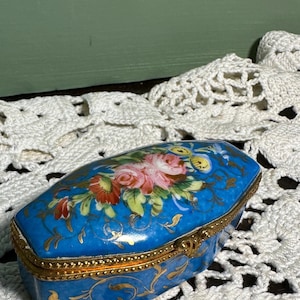 May include: A small, blue porcelain trinket box with a gold trim and floral design. The box has a hinged lid and is decorated with a bouquet of pink and red roses.