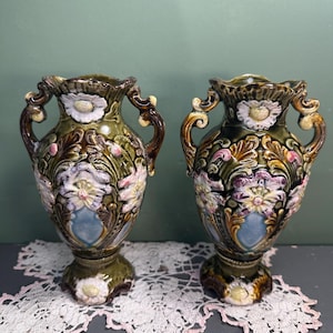 May include: Two antique ceramic vases with ornate floral designs. The vases are olive green with brown, white, pink, and blue accents. Each vase has handles and a pedestal base, and they are displayed on a lace doily.