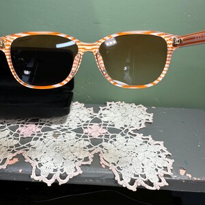 Puede incluir: Gafas de sol con montura a rayas naranjas y blancas y lentes marrones. Las gafas de sol tienen forma rectangular. El nombre de la marca es visible en la montura. El fondo es verde.