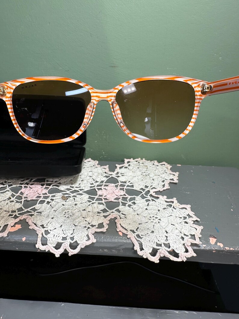 Puede incluir: Gafas de sol con montura a rayas naranjas y blancas y lentes marrones. Las gafas de sol tienen forma rectangular. El nombre de la marca es visible en la montura. El fondo es verde.