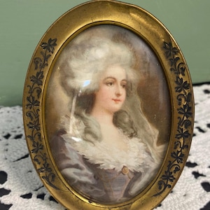 May include: Oval portrait of a woman in a gold frame. The portrait features a woman with white hair and a light blue dress with a white collar. The gold frame has a floral design around the edge.