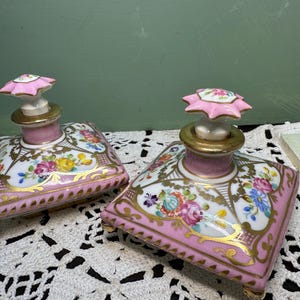 May include: Two antique porcelain perfume bottles. Each bottle is square with a pink base and floral design. The stoppers are pink and white, with gold accents. The bottles are on a white lace surface.