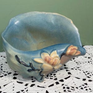 Pode incluir: Uma tigela de cerâmica azul com um design floral. A tigela tem uma forma curva com um exterior azul claro e um interior creme. O design floral apresenta flores creme e laranja com caules azuis escuros.