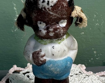 May include: A vintage ceramic doll with a dark complexion, wearing a white shirt with a green collar, blue shorts, and brown shoes. The doll has black hair with two side pigtails and a top knot. The doll is standing on a white lace doily.