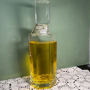 May include: A vintage glass bottle of Jean-Louis Scherrer perfume. The bottle is clear and filled with a yellow liquid. The label reads "Jean-Louis Scherrer Paris" and "Parfum".