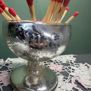 May include: A silver-toned, footed glass bowl filled with red-tipped matches. The bowl has an etched design and sits on a round base. The matches are arranged to be visible from the top. The background is a muted green.