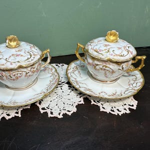 May include: Two vintage porcelain soup tureens with lids, featuring delicate floral and gold accents. The tureens are displayed on white lace doilies.