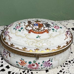 Könnte beinhalten: Ovale Schmuckdose aus Porzellan mit weißem Grund, verziert mit bunten Blumenmustern und goldenen Verzierungen. Der Deckel zeigt ein Wappen mit Löwe, Vogel und Banner mit Text. Wahrscheinlich Vintage oder antik.