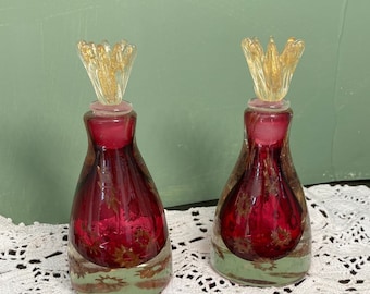 May include: Two vintage glass perfume bottles with red and gold accents. The bottles have a clear base and a gold-colored stopper. The bottles are displayed on a white lace doily against a green background.