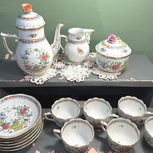 May include: A vintage porcelain tea set with floral designs. The set includes a teapot, creamer, sugar bowl, plates, and teacups. The teapot is topped with a rose. The plates and cups have gold trim.