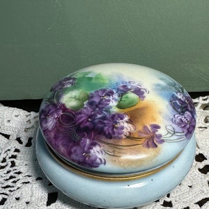 May include: A vintage porcelain trinket box with a light blue base and a lid decorated with a floral design of purple and white flowers on a green and yellow background.
