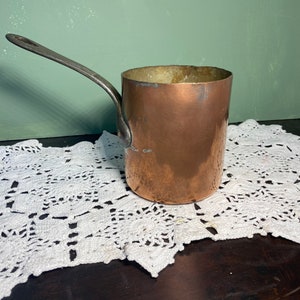 May include: A copper saucepan with a long, silver-colored handle. The pot has a cylindrical shape and shows signs of wear. The background is a muted green, and the pot rests on a white, lacy doily.