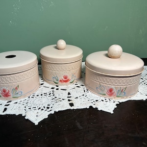 May include: Three vintage pink metal canisters with floral designs and wooden knobs. The canisters have a woven pattern and are decorated with pink roses and green leaves.