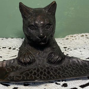 Pode incluir: Uma estatueta de gato de bronze empoleirada em cima de um prato em forma de peixe. O gato tem detalhes de pêlo e traços faciais, com as patas apoiadas no peixe. O peixe tem escamas e barbatanas, criando uma peça decorativa única.