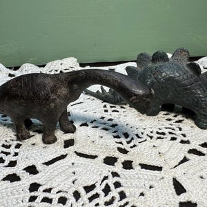 Puede incluir: Dos figuritas de dinosaurio negras, una con cuello largo y la otra con la espalda espinosa, están colocadas sobre una superficie de crochet blanco.