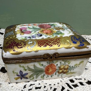 May include: A rectangular porcelain trinket box with a hinged lid. The box is white with floral and gold accents. The lid features a colorful floral design with gold trim. The box is closed with a metal clasp.