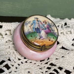 Puede incluir: Una pequeña caja de baratijas redonda y rosa con un borde dorado. La tapa presenta una escena pintada de dos figuras con ropa de época. La caja está sobre una superficie blanca de ganchillo.