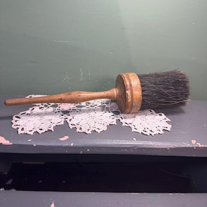 Könnte beinhalten: Eine antike Holzbürste mit dunklen Borsten. Die Bürste hat einen runden Kopf und einen langen, schlanken Griff. Die Bürste liegt auf einer weißen Spitzendecke. Der Hintergrund ist eine dunkelgraue Oberfläche.