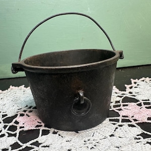 May include: A small, black cast iron pot with a handle. The pot has a worn, rustic look and is sitting on a white crocheted doily.