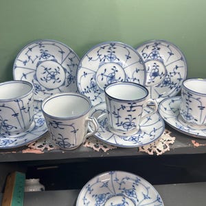 May include: A collection of white porcelain teacups, saucers, and plates with a blue floral design. The teacups have handles and are paired with matching saucers. The plates feature a similar blue pattern. The set is arranged on a dark surface.