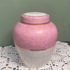 May include: A pink and white ceramic jar with a lid. The top half is pink with a gold floral pattern, and the bottom half is white with a textured design. Gold trim accents the jar and lid. The lid has small holes.