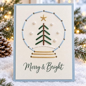 Könnte beinhalten: Eine rechteckige Grußkarte mit hellblauem Rand zeigt eine handgestickte Schneekugel mit einem grünen Weihnachtsbaum und einem goldenen Stern. Weiße Perlen akzentuieren das Design und der Text Merry & Bright ist auf der Karte aufgedruckt.