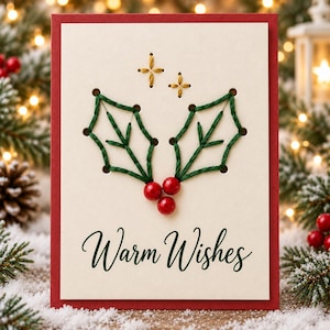 Puede incluir: Una tarjeta de felicitación rectangular con borde rojo y centro color crema. La tarjeta presenta un bordado en hilo verde que representa dos hojas de acebo, tres cuentas rojas como bayas y dos estrellas bordadas en oro. El texto dice Warm Wishes.